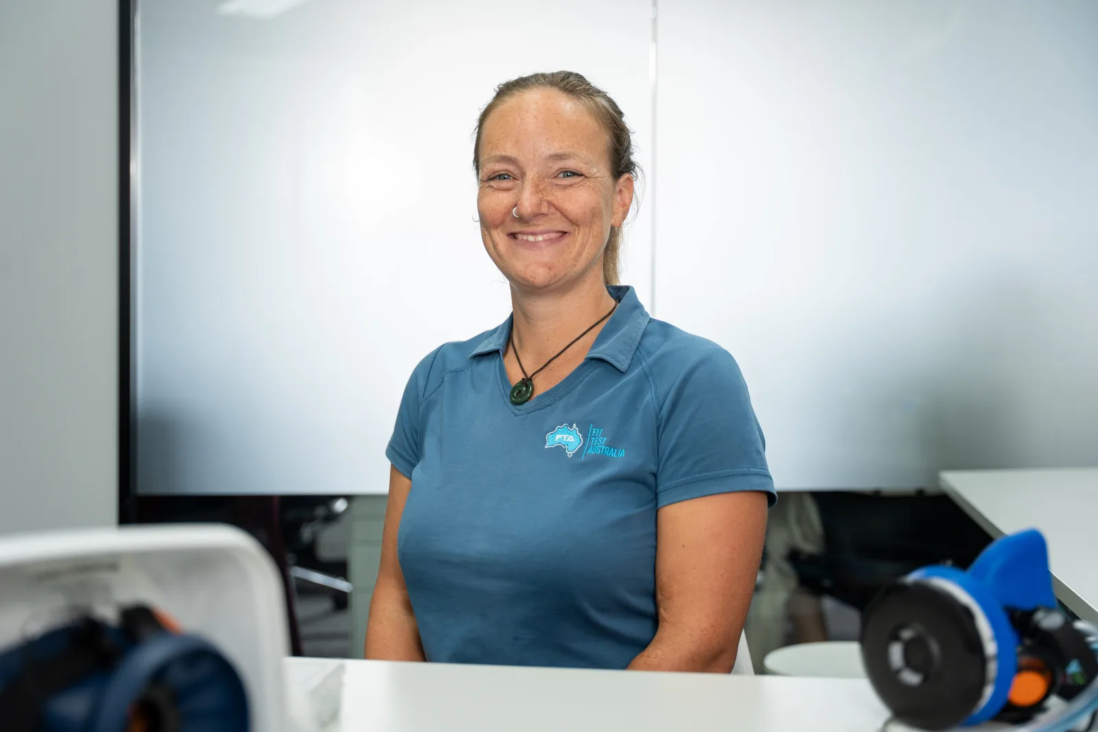 A Fit Test Australia fit-testing technician in the branded polo, respirators on the bench in front of her
